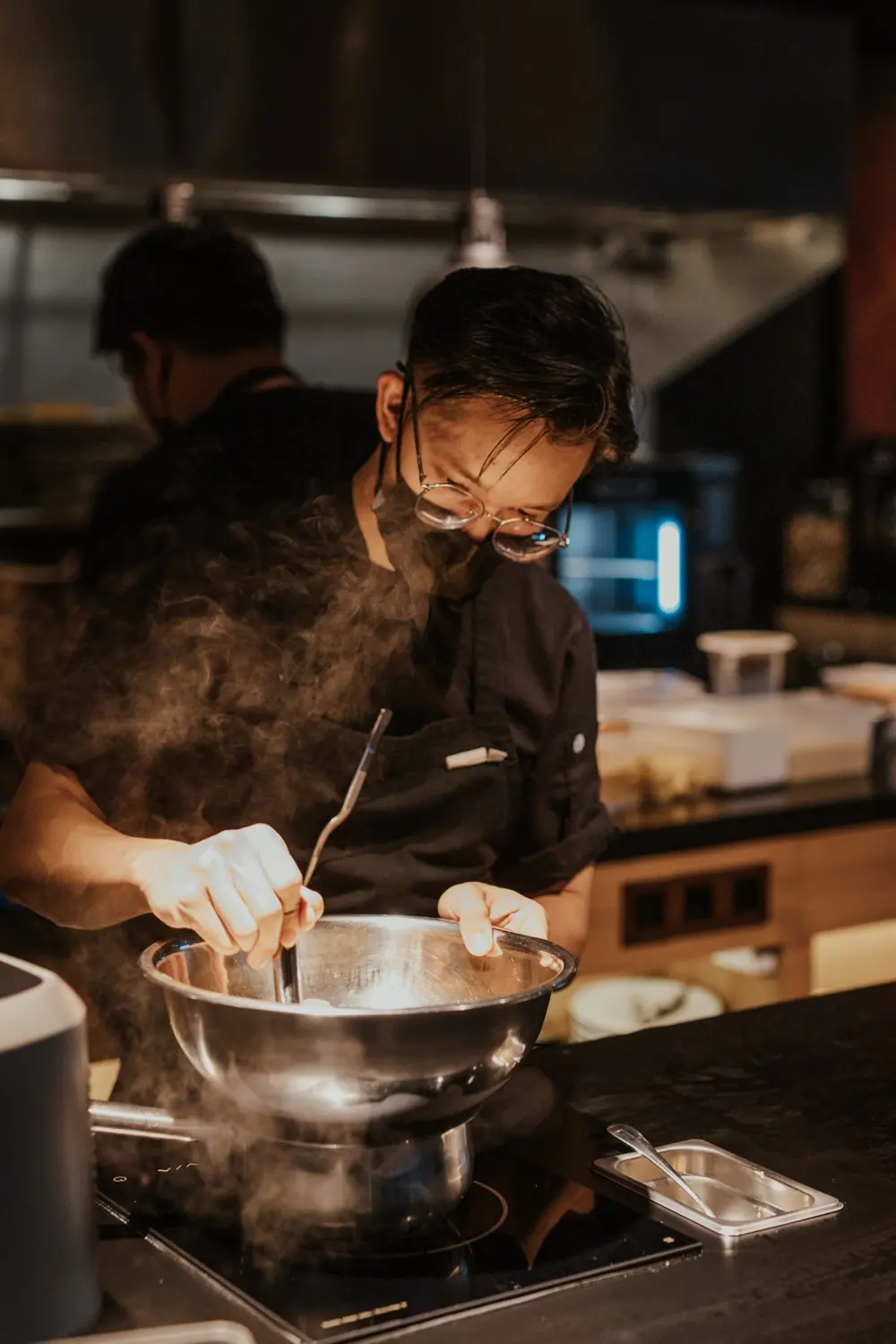 A chef in glasses and a face mask leans over a steaming pan at OITOM's open kitchen pass, a second cook working behind him.