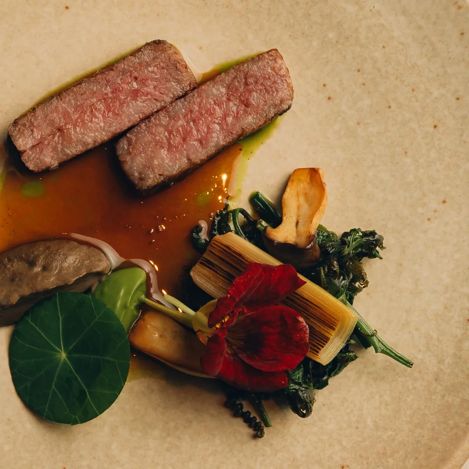 Two slices of rare seared beef in a glossy amber jus with grilled leek, mushrooms, wild greens, a nasturtium flower and leaf, on a speckled sand-coloured plate.