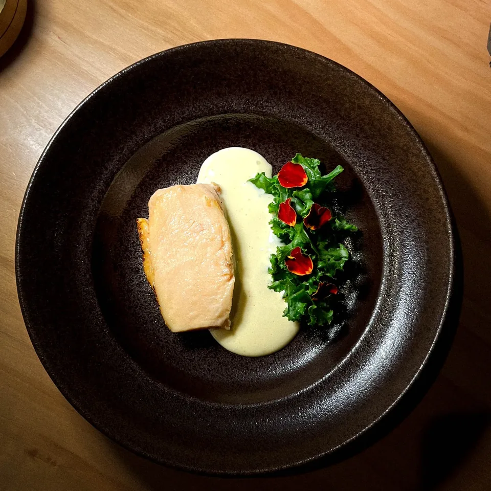 Overhead view of a seared fillet of fish with a pale butter sauce and curled kale scattered with red edible petals, in a dark glazed bowl on a pale wood table.